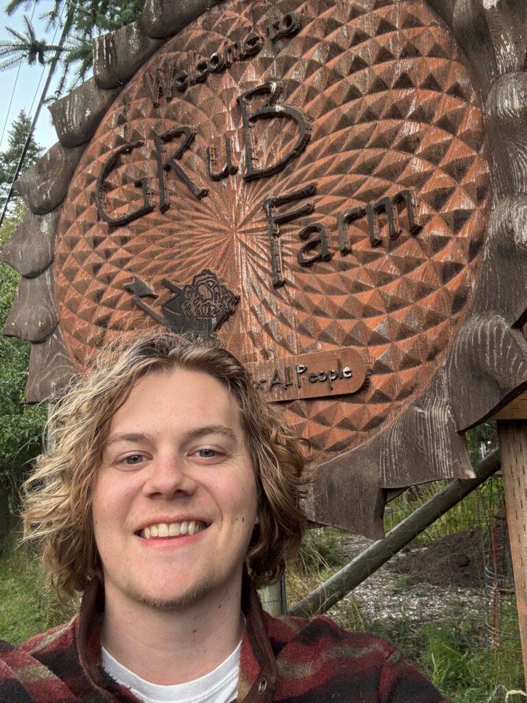 82EB1CC2-D690-4058-BE35-C3130C14A110 A person with wavy blond hair smiles in front of a large wooden sign that reads Welcome to GRuB Farm with decorative carvings and “All People” inscribed below. Green foliage appears in the background, celebrating the 2025 Day of Purpose.