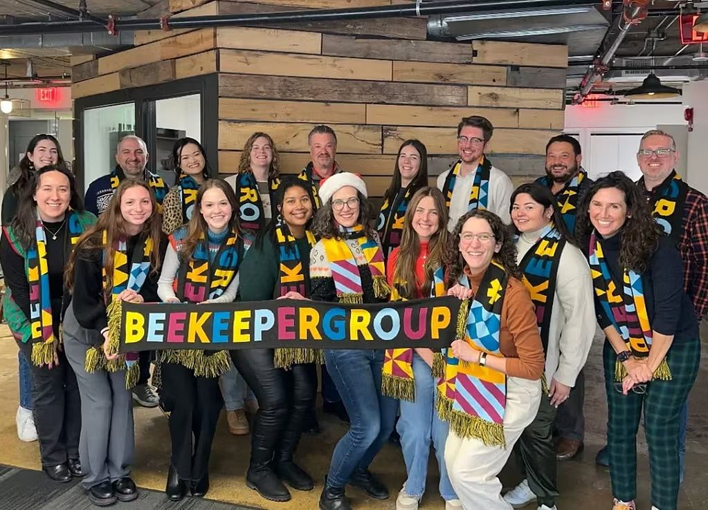about-group A group of people indoors pose and smile for a photo, each wearing a colorful scarf that spells out BEEKEEPER GROUP.